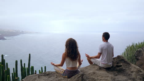 Seated-on-a-mountaintop,-a-man-and-a-woman-meditate-on-rocks,-hands-lifted,-as-they-take-in-the-ocean-and-engage-in-restful-breathing